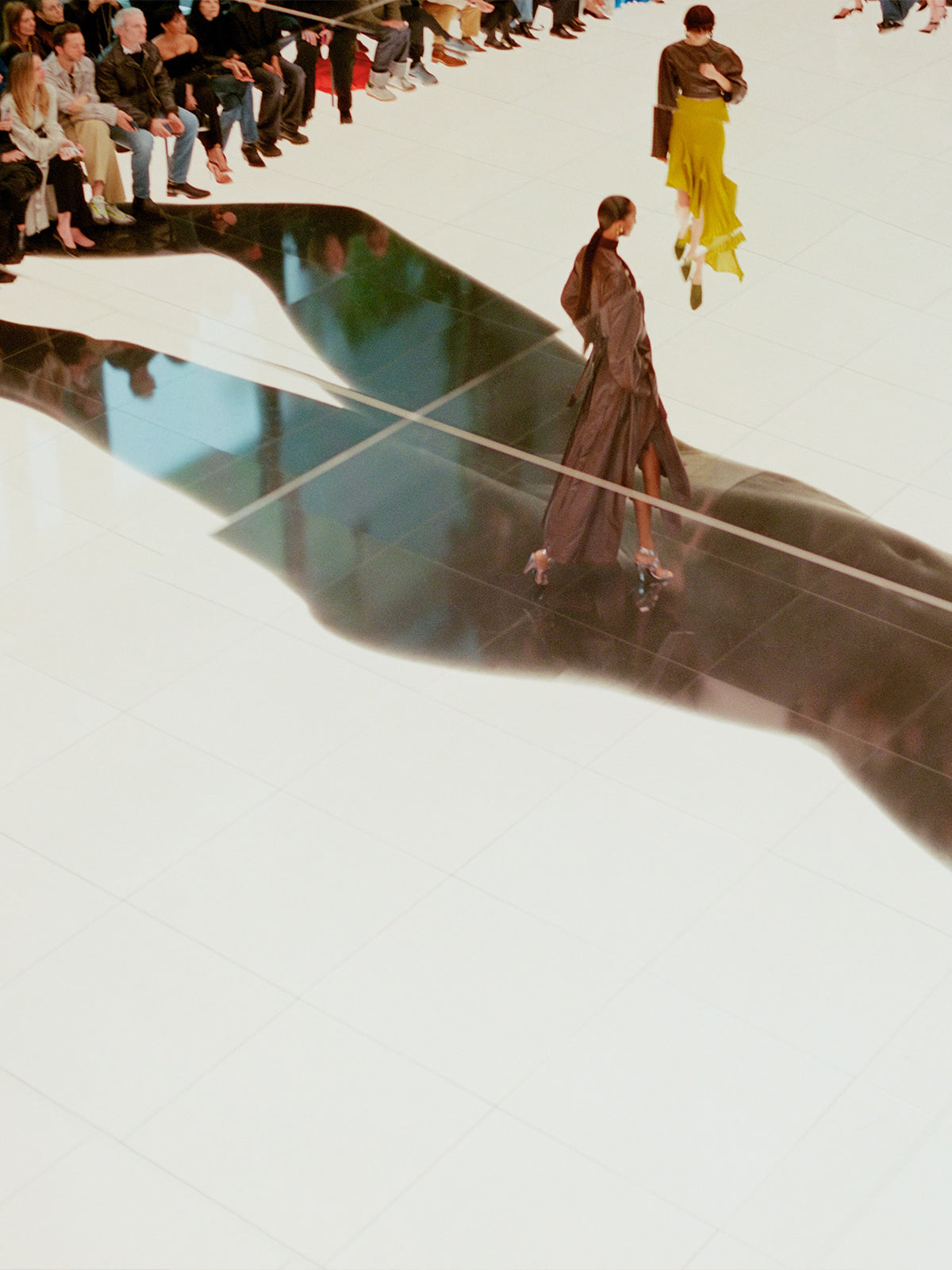 Woman walking on a reflective surface with a blurred crowd in the background at the Alaïa fashion show.