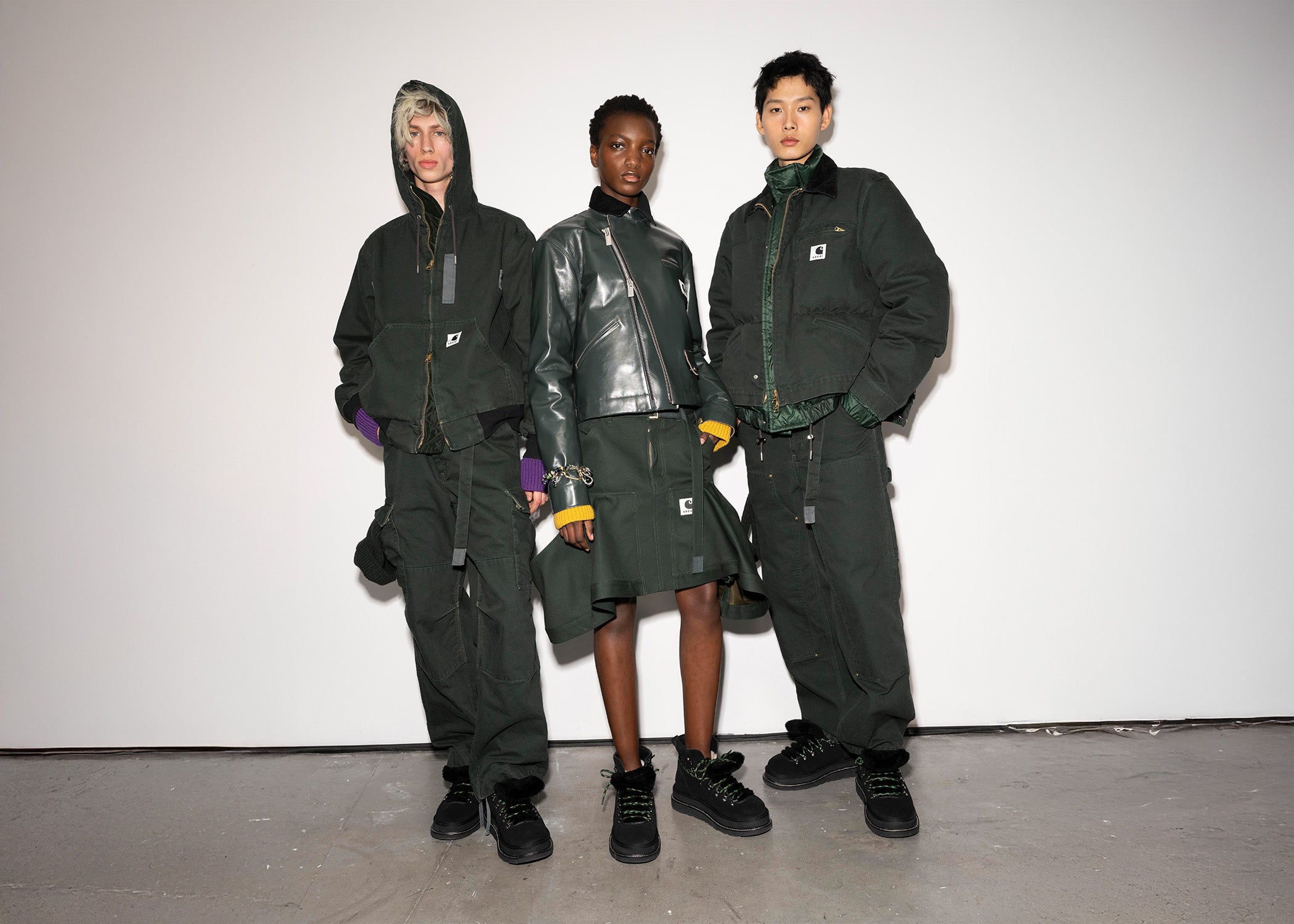 Three models showcase the Sacai x Carhartt WIP Autumn/Winter 2025 collection. They wear coordinated dark green utilitarian outfits with workwear-inspired details, including bomber jackets, cargo pants, and a leather jacket with a matching skirt. Each look is styled with layered elements, bright knit cuffs, and black lace-up boots. The models stand against a plain white backdrop, highlighting the collection’s structured silhouettes and rugged aesthetic.
