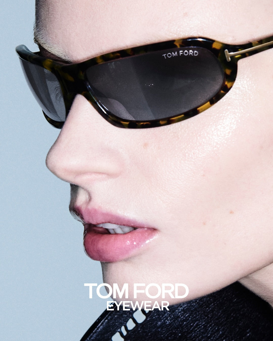 Close-up side profile of a model wearing Tom Ford sunglasses with a dark tortoiseshell frame and dark lenses, with the brand name visible on the upper frame and a Tom Ford Eyewear logo at the bottom.