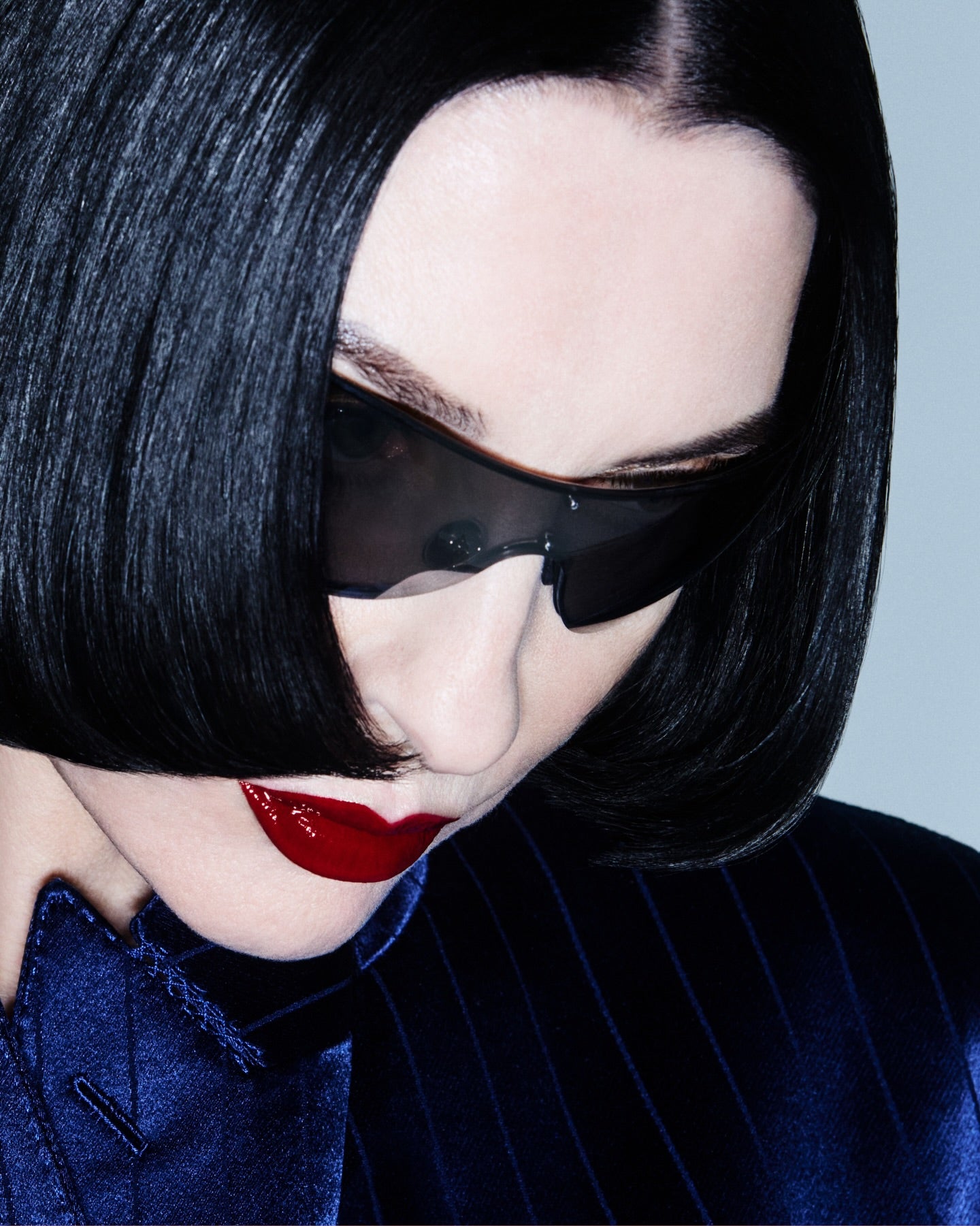 A close-up portrait of a woman with a dark bob haircut wearing dark tinted sunglasses, a dark blue pinstripe jacket, and bright red lipstick against a pale blue-gray background.