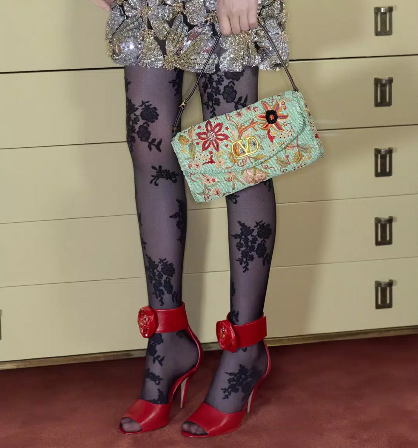 Person wearing Valentino floral tights and red high-heeled shoes, holding a colorful handbag.