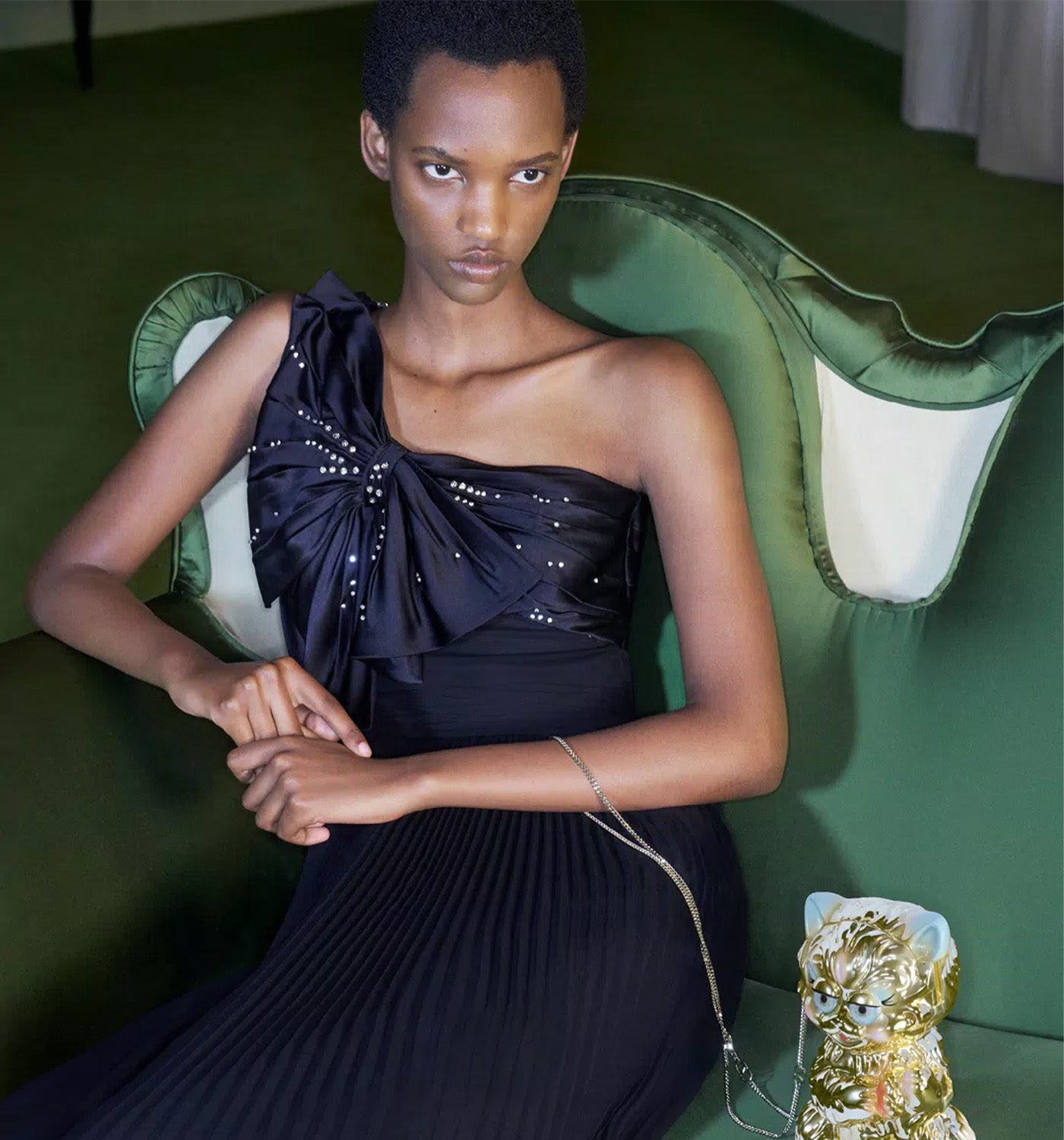 Woman in a Valentino black dress sitting on a green chair with a decorative vase on a table.