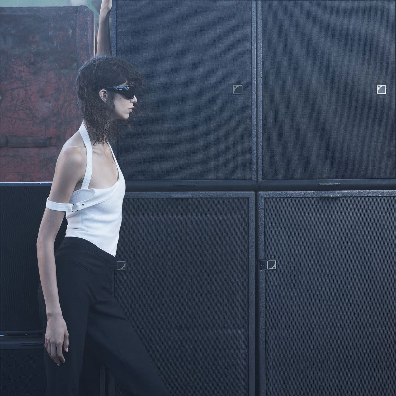 Woman in a white top and black pants standing against a dark wall.