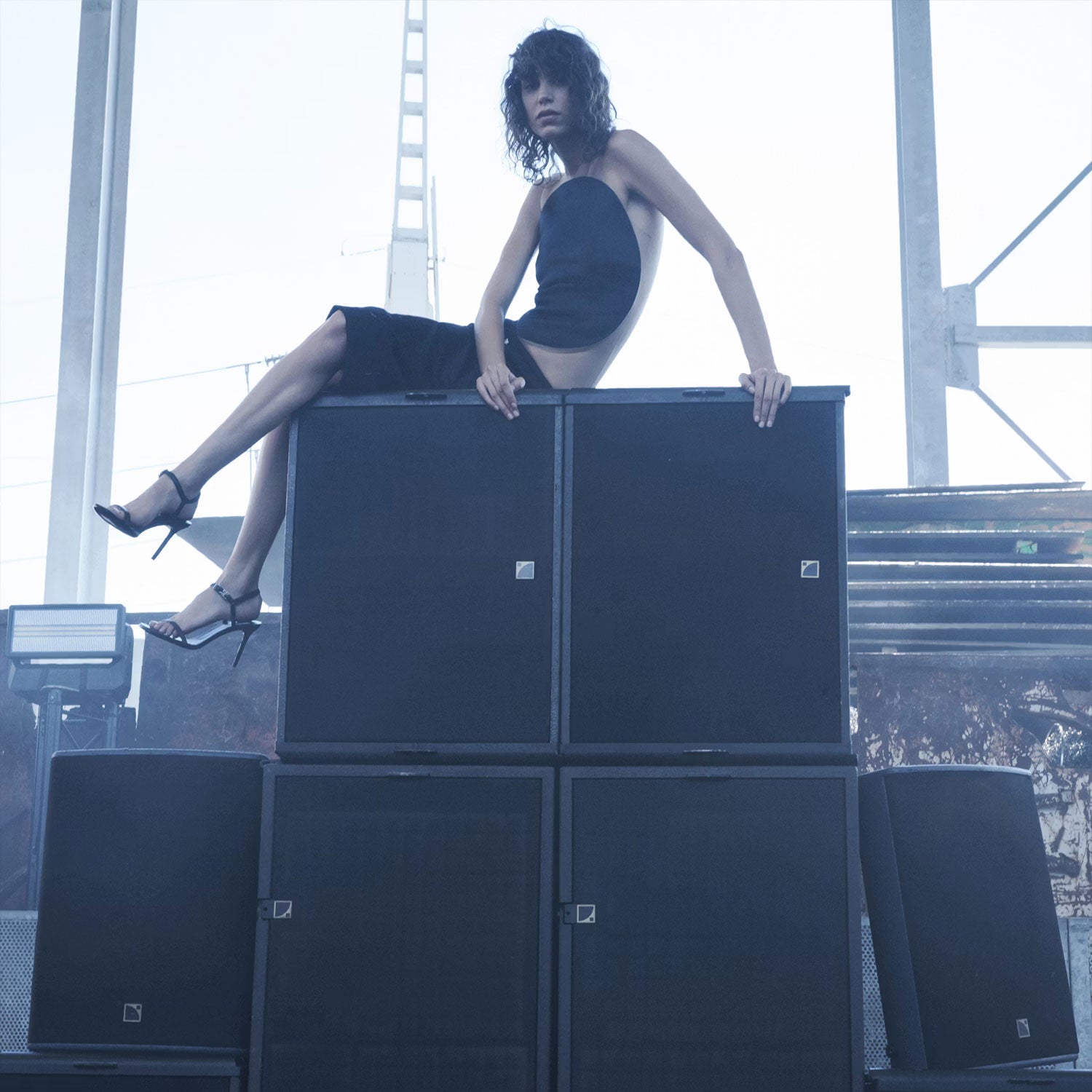 Woman in a black dress sitting on a stack of speakers with a blurred background